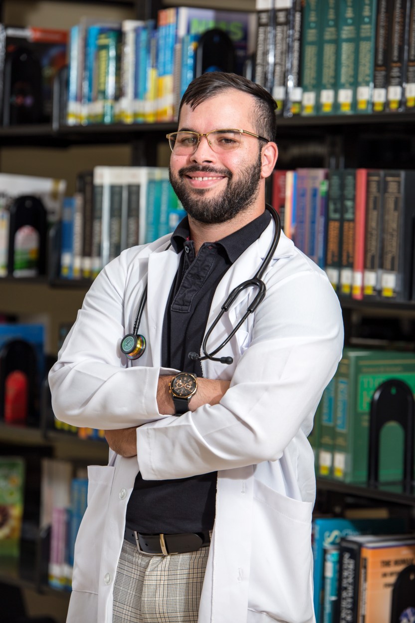 MUA Student Standing in Front of a Library, Wearing a White Coat and Stethoscope