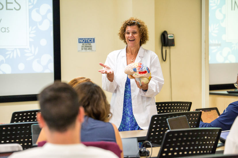 A woman in a lab coat presenting to a group of people during an MUA Faculty Lecture.